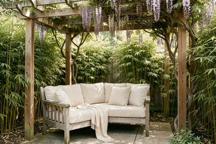 Coin ombragé aménagé sur une terrasse en été avec un lit de repos en teck et des coussins en lin crème sous une pergola couverte de glycine