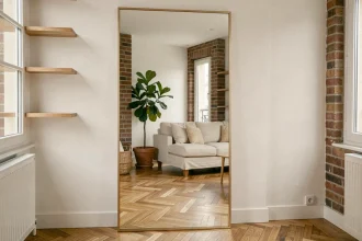 Grand miroir XXL posé contre un mur blanc dans un petit salon parisien, créant une illusion d'espace avec reflet d'un canapé en lin et parquet en chevrons